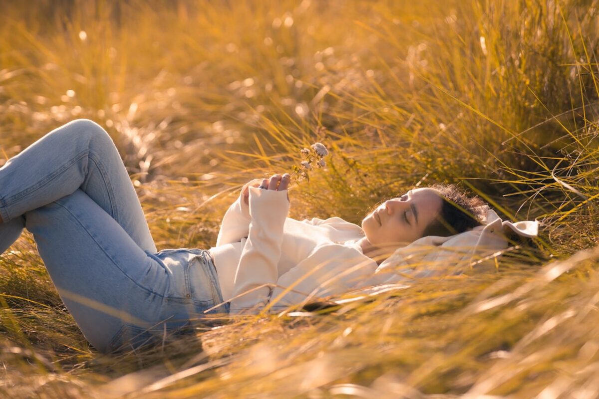 A woman in casual attire lies peacefully in a sunny grass field, embracing nature.