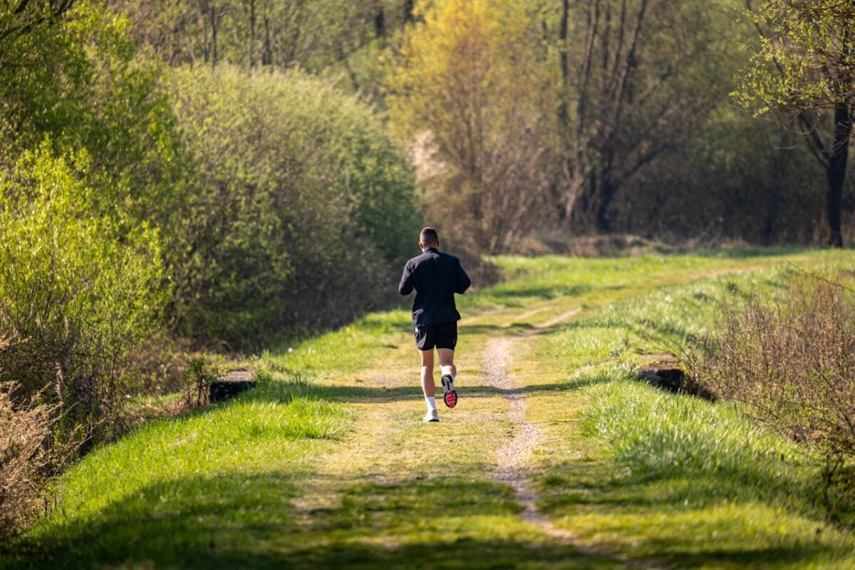 A man jogs on a scenic, grassy forest path surrounded by lush greenery under daylight.
