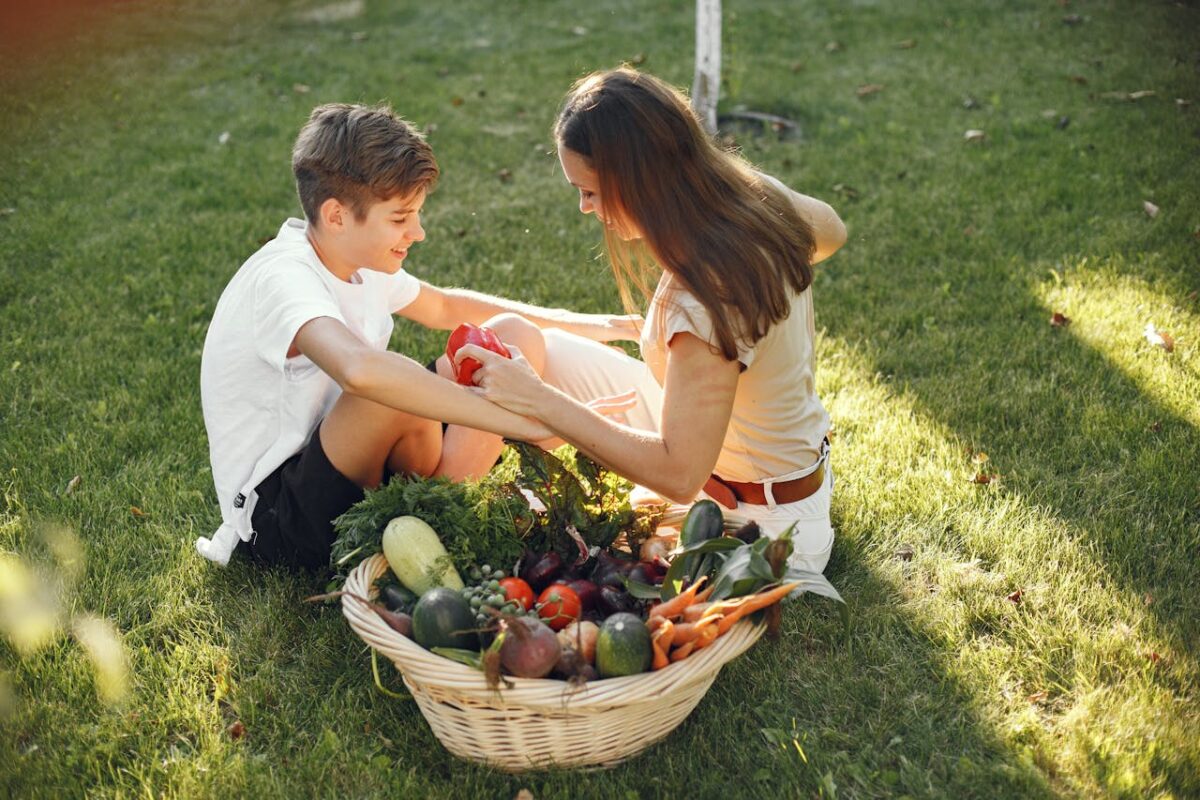 A mother and son sitting together on grass with fresh vegetables in a basket outdoors.