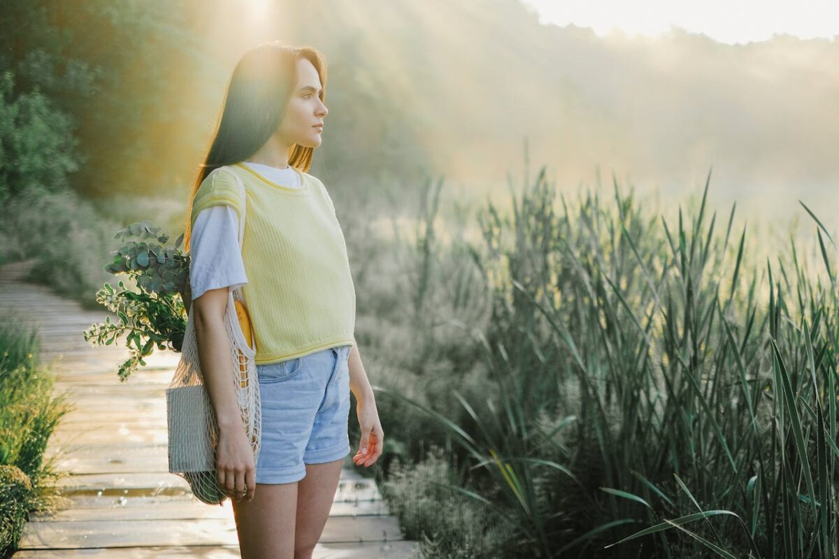 Woman wearing a yellow shirt and denim shorts standing on a sunny wooden pathway in nature.